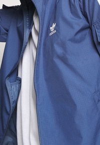 Blå lättviktsjacka med dragkedja fram, med en vit logotyp på vänster bröst, texturerat tyg och flera fickor.