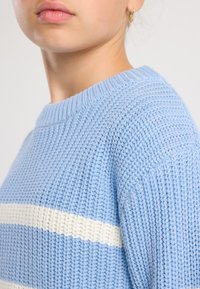 Close-up of a person wearing a light blue knitted sweater with a white horizontal stripe and a small gold earring.