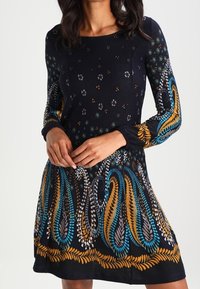 Långärmad klänning i mörk marinblå med blommiga och paisleymönster i turkos, guld och grått. Har en åtsittande bodice och en utsvängd kjol.