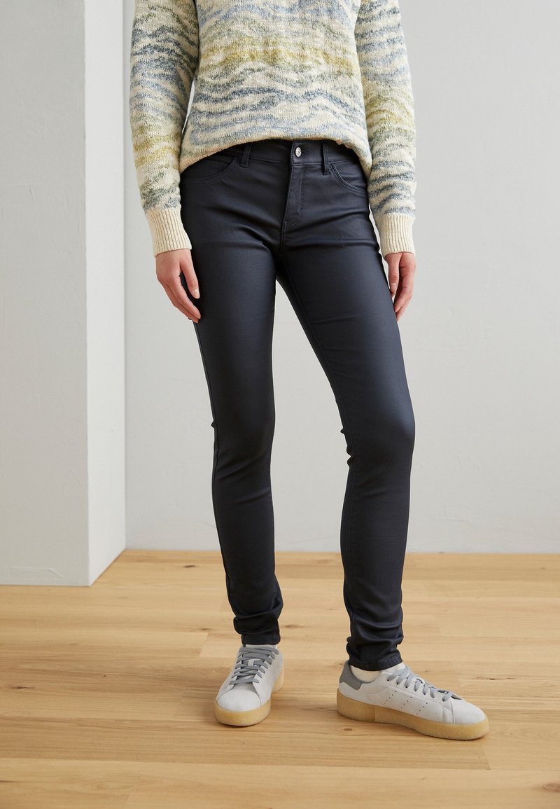 Jean noir à coupe slim, avec poches avant, braguette zippée et fermeture par bouton, porté avec des baskets gris clair sur un sol en bois.