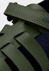 Žalios gumos sandalai su susikertančiomis dirželio juostomis, tekstūruota paviršiaus struktūra ir mėlynu akcentu dirželyje. Medžiagos artimas vaizdas.