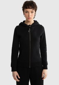 Młoda osoba o krótkich kręconych włosach nosi czarną bluzę z kapturem zapinaną na zamek błyskawiczny, z przednimi kieszeniami i regulowanym kapturem, stojąc na tle jednolitego jasnoszarego tła.