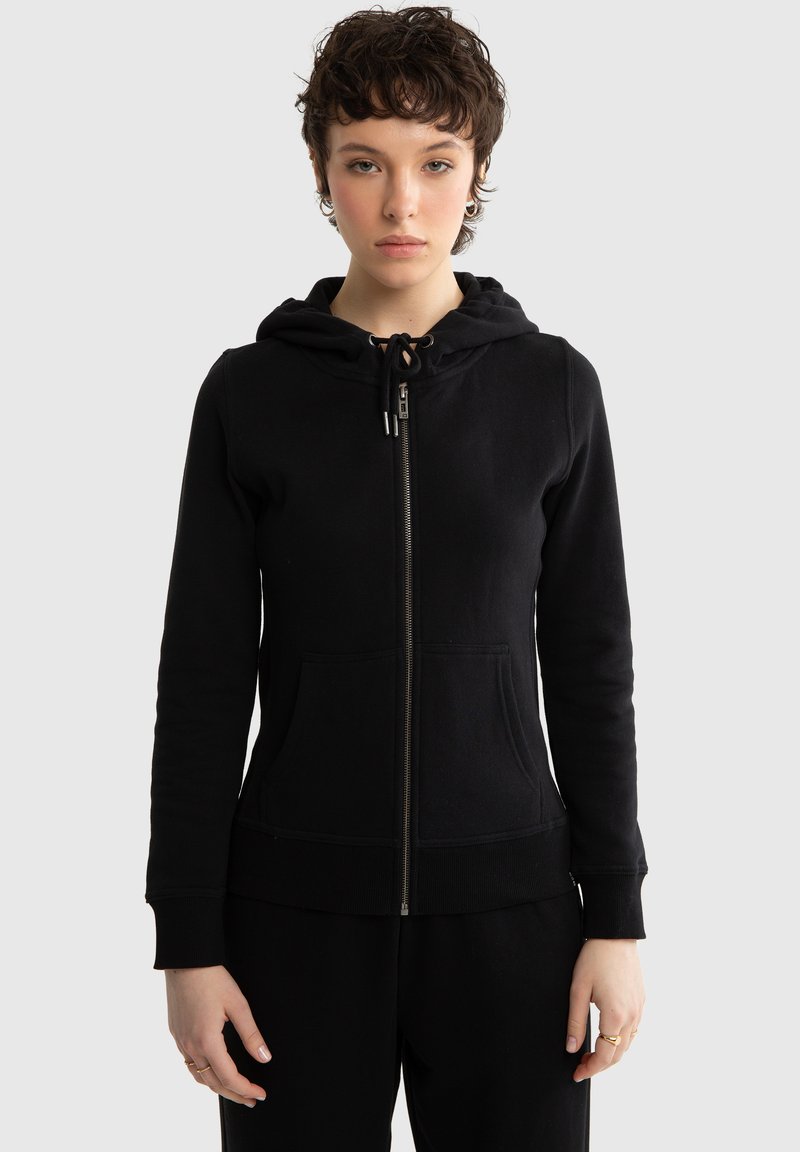 Młoda osoba o krótkich kręconych włosach nosi czarną bluzę z kapturem zapinaną na zamek błyskawiczny, z przednimi kieszeniami i regulowanym kapturem, stojąc na tle jednolitego jasnoszarego tła.