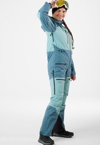 Ski suit in teal and light blue, featuring a fitted design, zippered pockets, adjustable cuffs, and boot gaiters, worn with goggles.