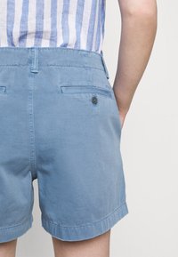 Blå shorts har ett slätt, strukturerat tyg med rullad fåll, två bakfickor med knäppningar och en midjehöjd i normal nivå.
