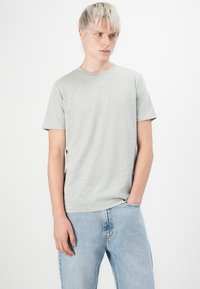 Persoană tânără cu părul blond scurt, îmbrăcată cu un tricou gri deschis și blugi albaștri deschis, stând cu o mână în buzunar, pe un fundal alb.