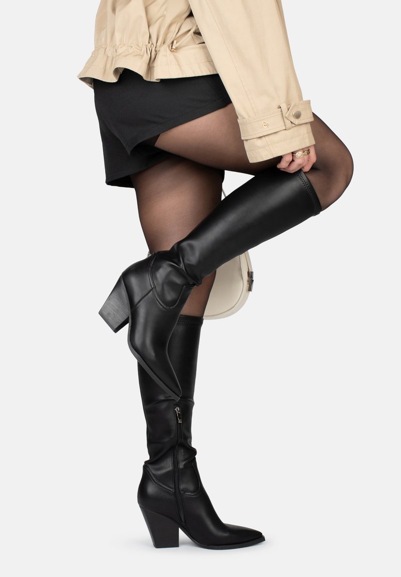 Botas negras de tacón alto hasta la rodilla con punta afilada, hechas de cuero suave. Presentan una cremallera lateral y un tacón apilado.