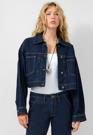 Frau mit langen blonden Haaren, die eine kurz geschnittene dunkle Jeansjacke, dazu passende Jeans, ein weißes Oberteil und eine große herzförmige Anhängerkette trägt.