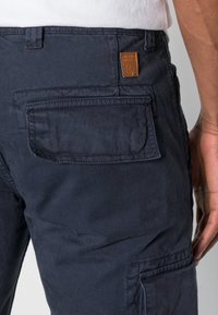 Námořnické nákladní kalhoty s texturovanou látkou, vybavené boční kapsou a malou hnědou koženou cedulkou s vyraženým nápisem „Alpha Industries“.