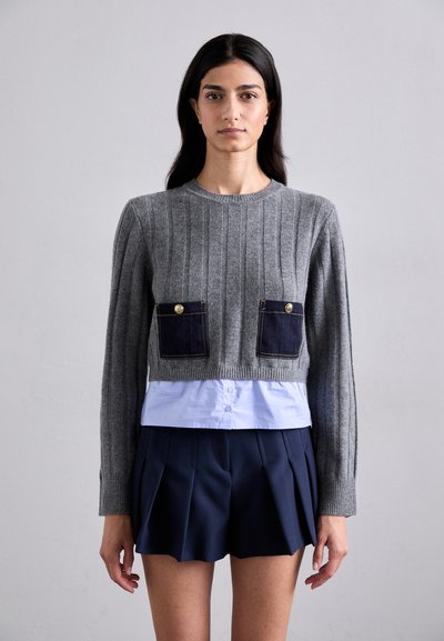 Suéter corto de punto de canalé gris con dos bolsillos de denim, que cuenta con botones dorados. Lleva encima de una camisa azul clara y pantalones cortos de plisado azul marino.