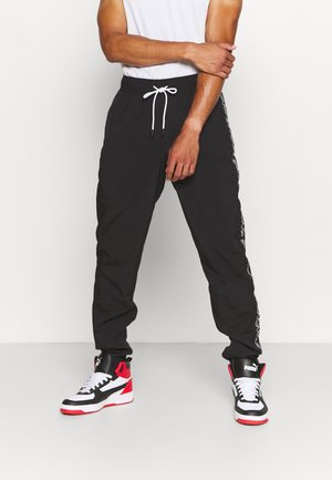 Mann trägt schwarze Jogger-Hosen mit weißem Kordelzug und schwarzen, weißen, roten High-Top-Puma-Sneakers, steht vor weißem Hintergrund.