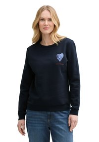 Sudadera de color azul marino en un material suave, con un gráfico de un corazón en el lado izquierdo del pecho y puños y dobladillo acanalados. Combinada con jeans azules.