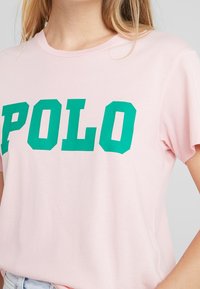 Růžové tričko s krátkým rukávem se silným zeleným nápisem "POLO", vyrobené z měkké textilie s volným střihem a klasickým kulatým výstřihem.