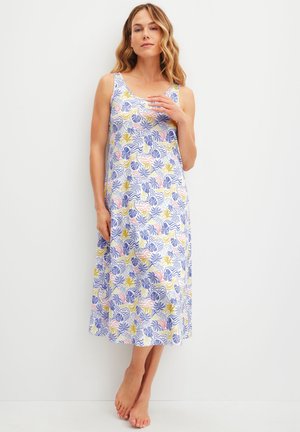 Vrouw staat blootsvoets en draagt een mouwloze, enkellange jurk met een meerkleurig patroon van blauwe, gele en roze bladeren.