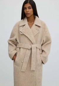 Žena v béžovém vlněném kabátu s páskem, širokými límci a kapsami, stojící před jednobarevným světlým pozadím.