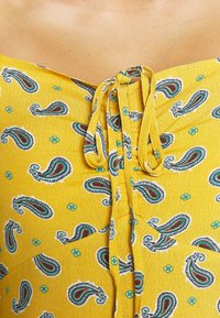 Keltainen kangastoppi, jossa on paisley-kuvio sinisen ja punaisen sävyissä sekä pieniä kukkakoristeita. Kauluksessa on solmittava yksityiskohta.