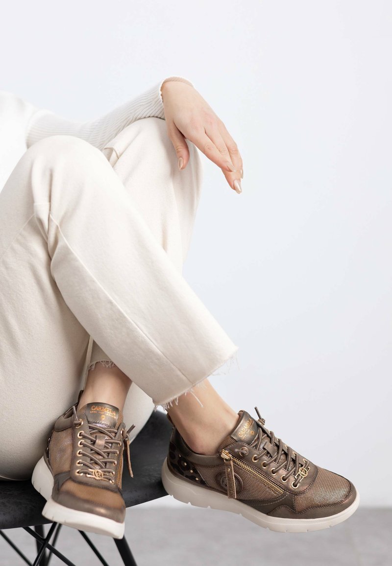Zapatillas de bronce fabricadas con malla y cuero, con herrajes dorados, un zipper lateral y una suela blanca. Llevadas con pantalones de color claro.