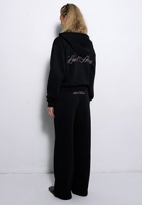 Czarna bluza z kapturem i spodnie dresowe w zestawie, charakteryzujące się luźnym krojem, miękką teksturą oraz napisem na plecach. Obie części mają elastyczne ściągacze.
