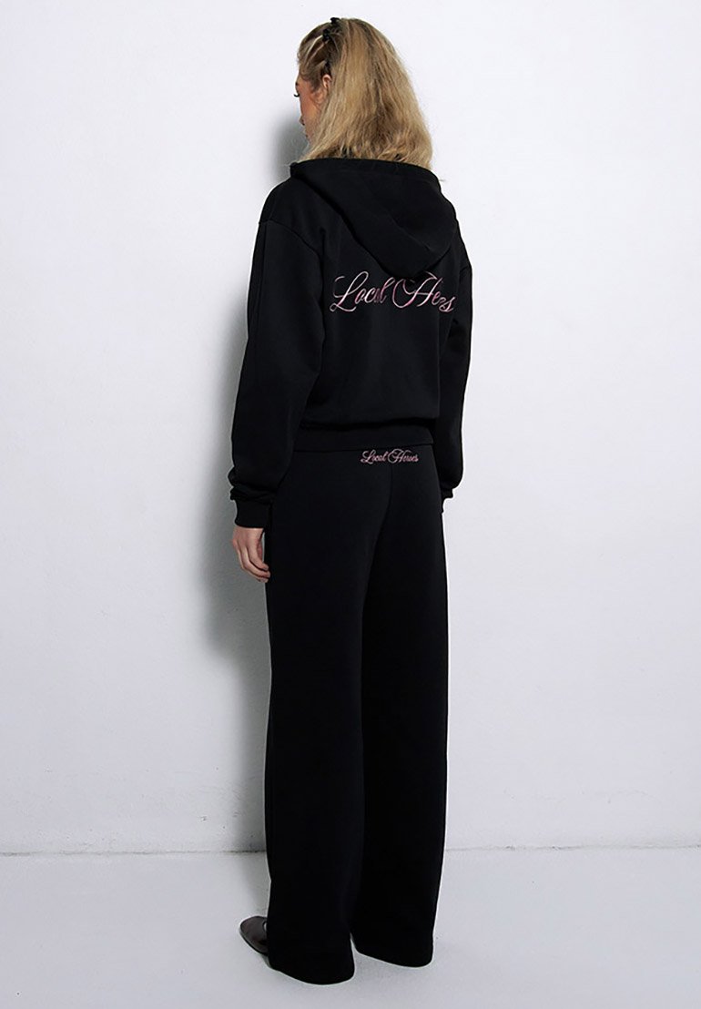 Czarna bluza z kapturem i spodnie dresowe w zestawie, charakteryzujące się luźnym krojem, miękką teksturą oraz napisem na plecach. Obie części mają elastyczne ściągacze.