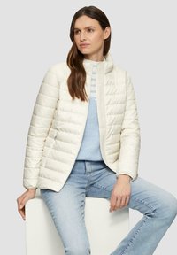 Veste matelassée crème avec col montant, fermeture éclair et deux poches latérales, associée à un haut en tricot bleu pastel et un jean bleu clair.