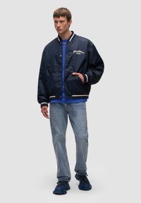 Granatowa kurtka bombowa z białymi paskami na mankietach i dolnym obwodzie, z wyszywanym logo. Jasnoniebieskie dżinsy i ciemne sneakersy dopełniają stylizację.