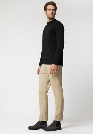 Czarny sweter z długim rękawem i wysokim kołnierzem, zestawiony z beżowymi spodniami i czarnymi botkami za kostkę. Gładkie tekstury w całym zestawie.