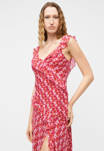 Γυναίκα με κυματιστά ξανθά μαλλιά φοράει αμάνικο φόρεμα με ροζ και κόκκινο σχέδιο, βολάν και σκίσιμο μέχρι το μηρό, ποζάροντας μπροστά σε απλό φόντο.
