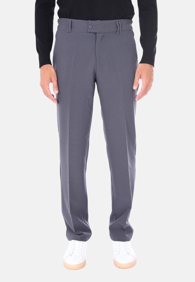 Uomo che indossa pantaloni grigi su misura, maglione nero a maniche lunghe e sneakers bianche, in piedi con le braccia rilassate ai fianchi su uno sfondo bianco.