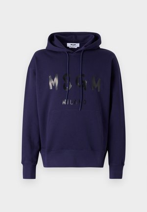 Henkilö, jolla on yllään laivastonsininen huppari, jossa teksti "MSGM Milano", yhdistettynä väljiin mustiin cargo-housuihin ja mustiin kenkiin yksiväristä taustaa vasten.