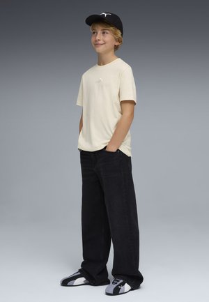 Niño con camiseta beige de Puma, gorra negra, pantalones anchos negros y zapatillas Puma a juego, de pie con las manos en los bolsillos, sonriendo levemente.