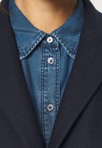 Donker marineblauwe blazer van textuurstof, gedragen over een lichtblauw denim shirt. Het shirt heeft een puntkraag en knoopdetails.