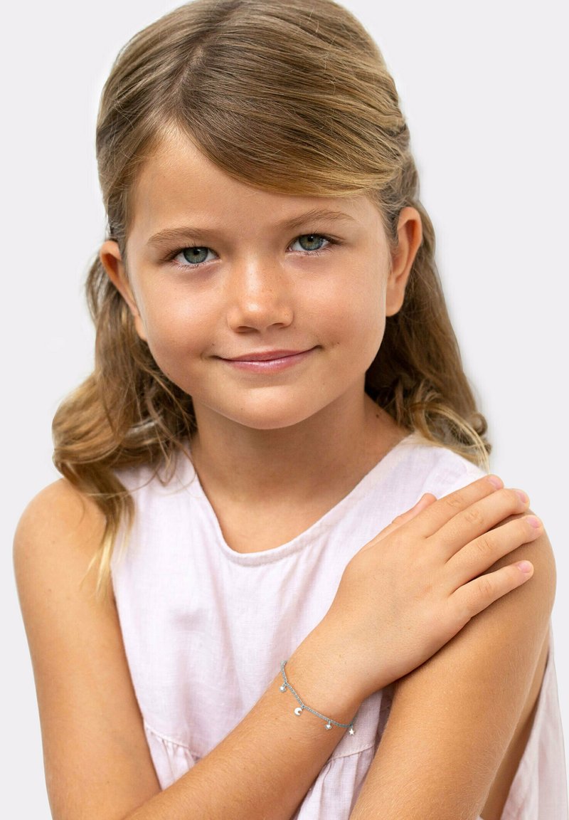 Bracciale in catena argento con piccoli charm a forma di luna e stella, indossato al polso di un bambino. Top senza maniche di un delicato viola e tono di pelle naturale.