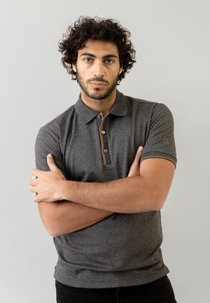 Polo grigio a maniche corte, con bottoni e dettagli a contrasto sul colletto, indossato da una persona con le braccia incrociate su uno sfondo chiaro.