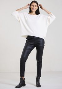 Weißer, oversized Pullover mit Dreiviertelärmeln, kombiniert mit schwarzer, enganliegender, Kunstlederhose und schwarzen Anklets.