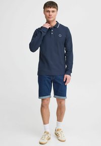Marine langermet polo-skjorte med hvit krage, med en liten logo-detalj på brystet, kombinert med denimshorts og joggesko.