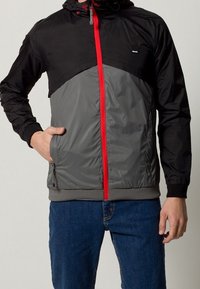 Svart och grå vindjacka med röd dragkedja, utrustad med huva, elastiska ärmslut och sidofickor. Slät yta, sportig design.