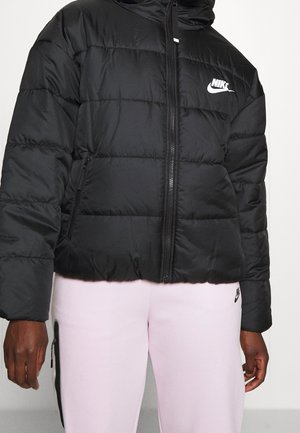 Czarna pikowana kurtka Nike z zamkiem z przodu i kieszeniami bocznymi, noszona z jasnoróżowymi spodniami dresowymi Nike z małym logo na udzie.