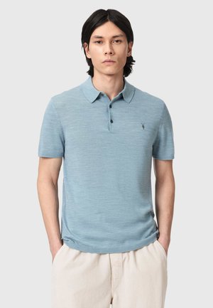 Ung mand med glat mørkt hår iført en lyseblå kortærmet polo og lysebeige bukser, stående med hænderne i lommerne.