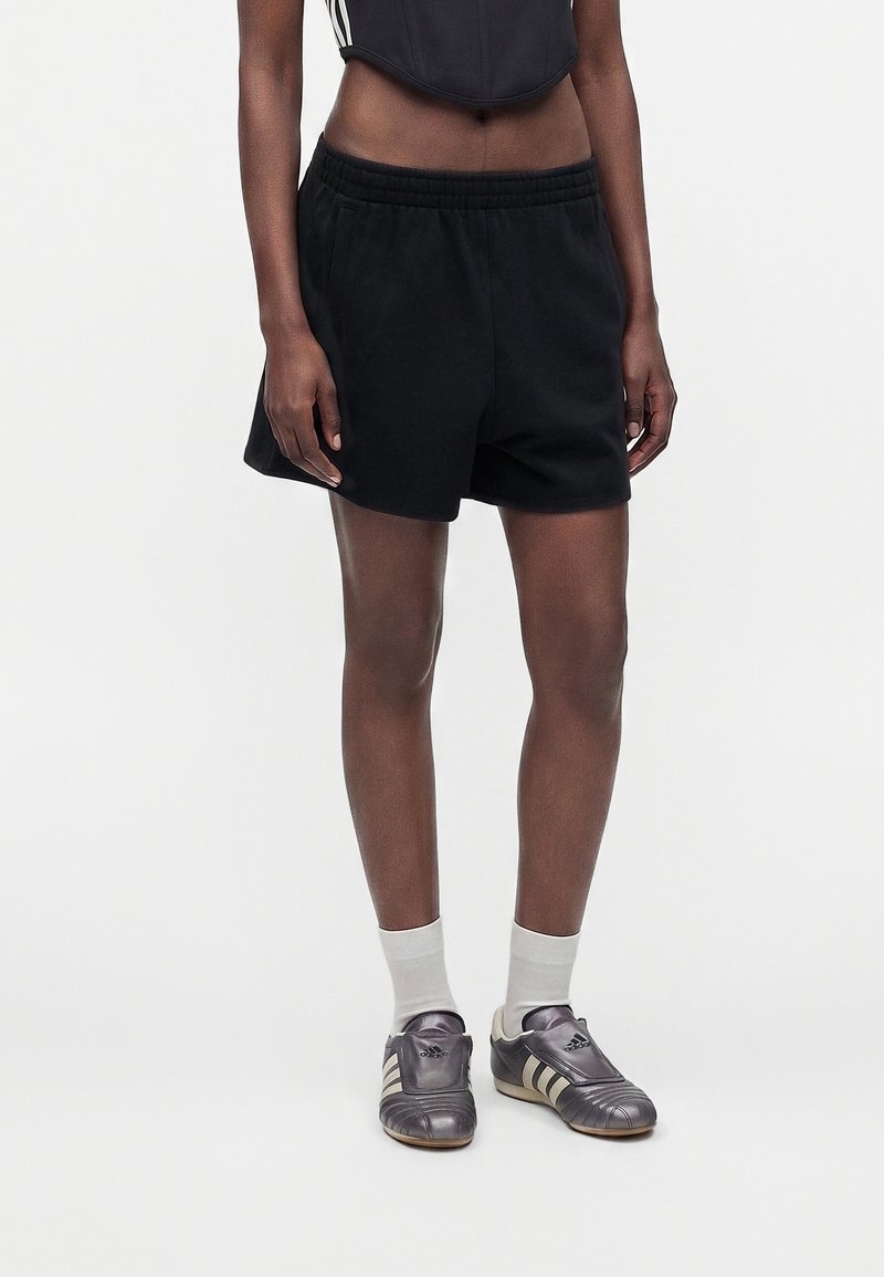 Personne portant un short noir, des chaussettes blanches et des baskets gris métallisé, debout devant un fond clair uni.