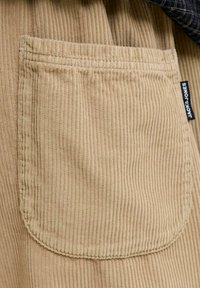 Poche en tissu velours côtelé beige avec des rainures verticales et une étiquette noire "JACK & JONES" sur le côté d'un vêtement.