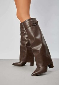 Bottes en cuir marron mi-mollet au design légèrement tombant, avec une accent en boucle en haut et un bout pointu sur un talon bloc.