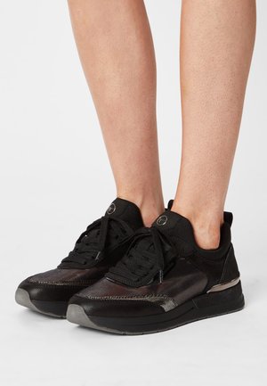 Black low-top sneakers with textured design and rounded toe worn on bare legs, shown against a plain white background.