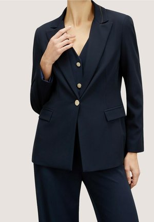 Femme portant un blazer bleu marine foncé sur mesure avec trois boutons dorés et un pantalon assorti, posant avec une main près du col et l'autre le long du corps.