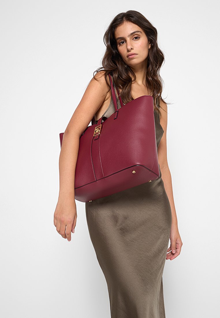 Bolso tote de cuero marrón oscuro con un diseño estructurado, asas superiores y acentos de hardware en tono dorado. Textura suave y forma rectangular.