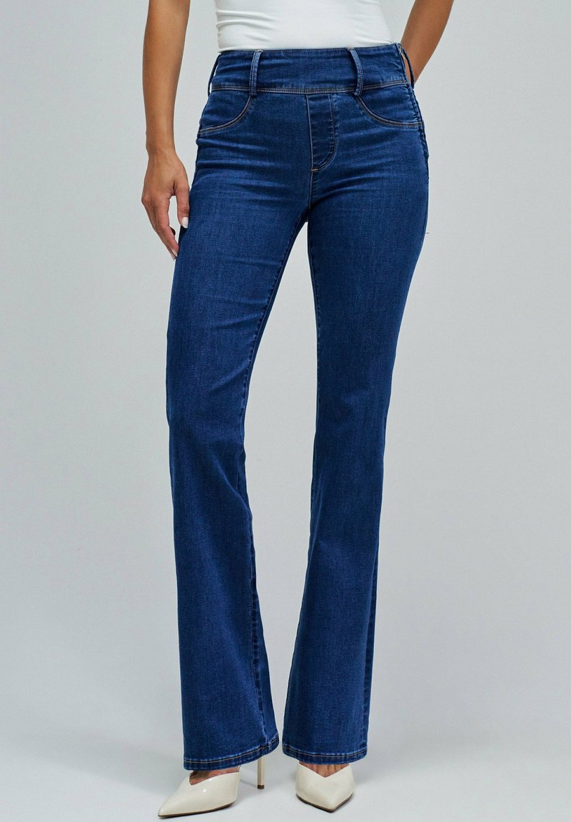 Jeans de mezclilla de color azul oscuro, acampanados, con una cintura de altura media, que presentan detalles de costura sutiles y una textura suave. Los zapatos son de punta y blancos.