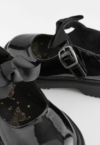 Chaussures Mary Jane en cuir verni noir avec brides à nœud en gros-grain, boucle argentée, semelles en caoutchouc, et semelles intérieures imprimées de papillons sur fond blanc.