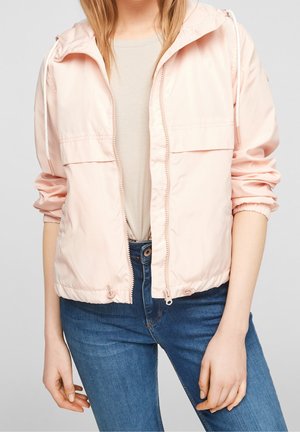 Person trägt eine hellrosa Kapuzenjacke mit Reißverschluss über einem beigefarbenen Hemd, kombiniert mit blauen Jeans, steht vor einem einfarbigen Hintergrund.