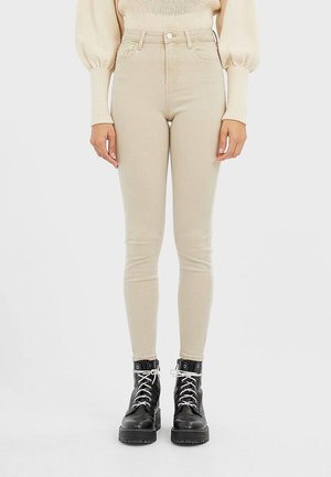 Personne portant un pantalon skinny beige taille haute, un pull en tricot crème avec des manches bouffantes, et des bottes de combat noires à lacets, debout devant un fond uni.