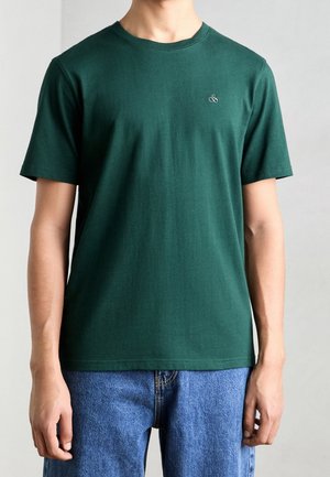 Uomo che indossa una maglietta verde scuro a maniche corte con piccolo logo bianco sul petto sinistro e jeans blu.