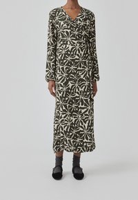 Langærmet omslagskjole i mørkegrøn med et hvidt botanisk mønster. Har en dyb V-udskæring og falder under knæet, parret med sorte sko.
